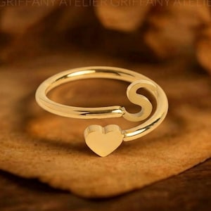 May include: A gold-toned open ring featuring a polished finish. One end of the band terminates in a stylized letter S, while the opposite end features a small, solid heart-shaped charm. The ring has a smooth, continuous metal band design.