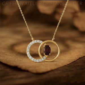 May include: A gold-colored necklace featuring two interlocking circles. One circle is encrusted with small, clear gemstones, while the other holds a deep red oval gemstone. The necklace hangs on a delicate gold chain.