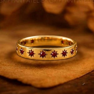 Op de afbeelding: Een gouden ring met een reeks gelijkmatig verdeelde stervormige zettingen, elk voorzien van een kleine, ronde, dieprode edelsteen. Het metalen oppervlak is gepolijst en glad, waarbij de stermotieven de gehele buitenomtrek sieren.