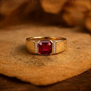 Könnte beinhalten: Ein Goldring mit einem quadratisch geschliffenen, tiefroten Edelstein in der Mitte, flankiert von zwei vertikalen Reihen kleiner, klarer runder Steine, die in die Ringschiene eingelassen sind. Der Ring hat eine glatte, polierte Oberfläche.