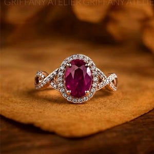 May include: An oval-cut deep red gemstone ring set in a rose-colored metal band. The central stone is surrounded by a halo of small white stones, with additional white stones embedded along the twisted, crisscrossing shank of the ring.