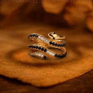 Op de afbeelding: Een goudkleurige ring in de vorm van een slang met een wikkelontwerp. Het lichaam van de slang is ingelegd met afwisselende rijen heldere witte stenen en kleine zwarte stenen, met een gladde gouden kop aan één uiteinde van de ring.