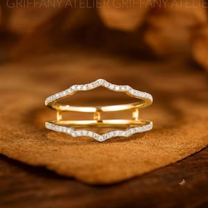 May include: A gold ring guard featuring two parallel bands connected by vertical bars. Both bands have a wavy, chevron-like shape and are encrusted with small, clear, sparkling stones along the outer edges, creating a double-layered design.