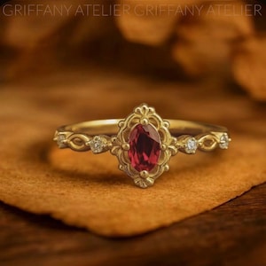 May include: A gold ring with a central oval-shaped, deep red gemstone. The ring features an ornate, filigree design around the gemstone and small accent diamonds. The band has a delicate, interwoven pattern. The ring is set against a warm, brown background.