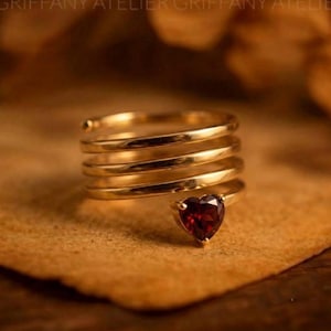 May include: A gold-toned spiral ring featuring four stacked bands that wrap around the finger. The end of the ring is adorned with a single, faceted, heart-shaped dark red gemstone set in a prong setting, creating a continuous coiled design.