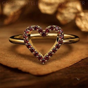 May include: A gold ring featuring a heart-shaped centerpiece outlined with small, round, deep red garnet gemstones. The band is smooth and polished, connecting to the base of the heart. The text GRIFFANY ATELIER is visible at the top of the image.