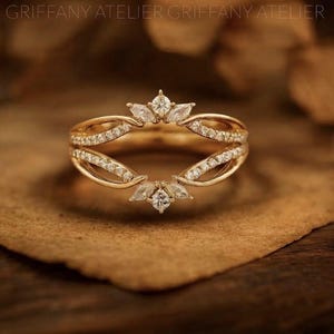 Könnte beinhalten: Ein goldener Ring-Guard mit geteiltem Banddesign, verziert mit kleinen runden Pavé-gefassten Steinen sowie marquise-geschliffenen Steinen an den oberen und unteren Mittelpunkten. Das Metall hat einen warmen Goldton und die Steine sind klar.