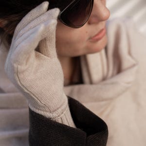 Op de afbeelding: Close-up van een lichtbeige gebreide handschoen. De handschoen wordt gedragen met een donkergrijze jas. De persoon draagt een zwarte zonnebril. De handschoen is gemaakt van een zacht, getextureerd materiaal en de manchet is geribbeld.