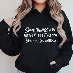 Puede incluir: Sudadera con capucha negra con bolsillo delantero y capucha con cordón. El texto blanco en la parte delantera dice: "Algunas cosas es mejor dejarlas solas, como yo, por ejemplo."