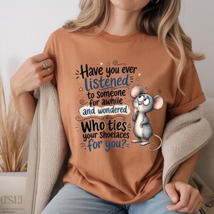May include: A rust-colored t-shirt featuring a cartoon mouse and the text: Have you ever LISTENED to someone for awhile and wondered WHO ties your shoelaces for you? The design uses black and blue lettering with decorative swirls and stars.