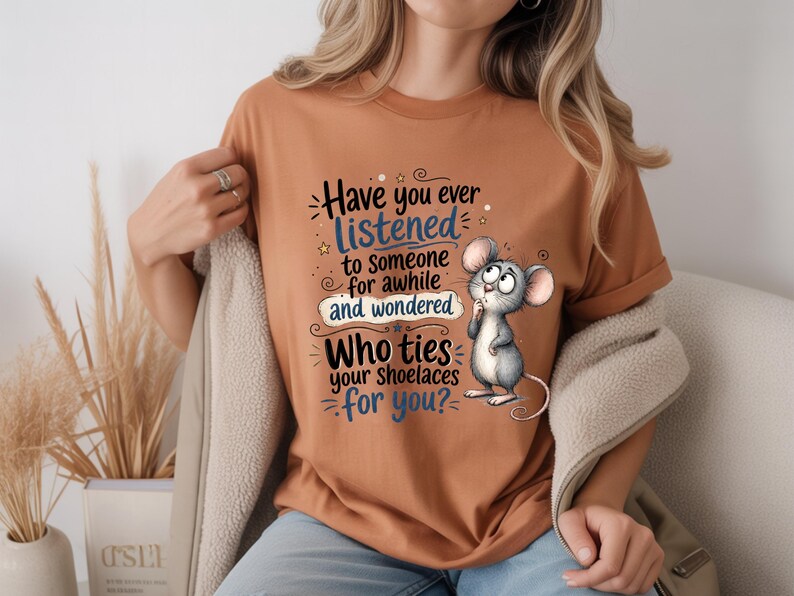 May include: A rust-colored t-shirt featuring a cartoon mouse and the text: Have you ever LISTENED to someone for awhile and wondered WHO ties your shoelaces for you? The design uses black and blue lettering with decorative swirls and stars.