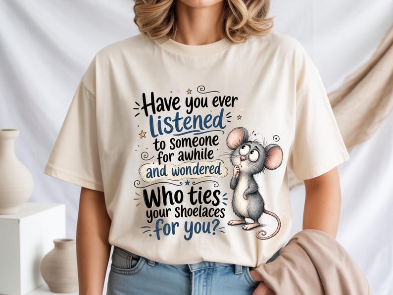 May include: A cream-colored t-shirt featuring a cartoon mouse with large ears and a thoughtful expression. The text reads: Have you ever listened to someone for awhile and wondered Who ties your shoelaces for you? The design uses black and blue ink.