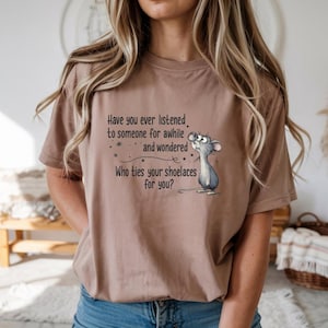 May include: A tan t-shirt featuring the text "Have you ever listened to someone for awhile and wondered Who ties your shoelaces for you?" along with a cartoon mouse illustration. The shirt is a neutral color.