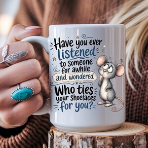 May include: A white ceramic mug featuring a cartoon illustration of a grey mouse and the text: Have you ever listened to someone for awhile and wondered Who ties your shoelaces for you? The mug has a handle and a smooth, glossy finish.