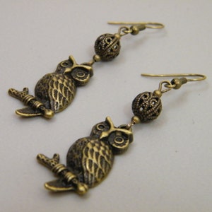 May include: A pair of bronze owl earrings with a filigree ball charm. The owls are perched on a branch and have detailed features.