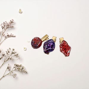 Peut inclure: Trois pinces à cheveux en résine rouge, violet et rouge avec des clips en métal doré. Les pinces ont un aspect scintillant et texturé. Le fond est blanc avec une branche de fleurs blanches.