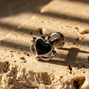 May include: A silver-colored ear piercing stud with a black heart-shaped center. The stud has a decorative design around the heart and a post for insertion. The jewelry is displayed on a textured, light brown surface.
