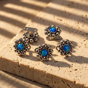 May include: Five silver-toned flower-shaped earrings with a central blue opal stone. Each earring features a cluster of small, round beads around the central stone. The earrings are displayed on a textured, light-colored surface.
