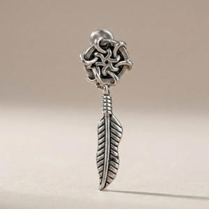 May include: Silver-toned earring featuring a decorative top with a woven design, connected to a detailed feather charm. The earring has a post for secure wear. The design is intricate and detailed, suitable for a variety of styles.