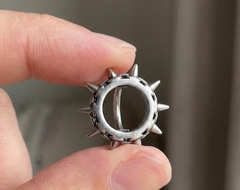 May include: A silver-colored ear cuff with a circular design and pointed spikes around the outer edge. The ear cuff is held between two fingers against a neutral background. The jewelry is a unique accessory.