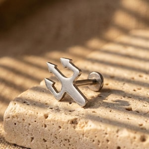 May include: A silver-colored trident-shaped earring stud. The three-pronged design is centered on a flat, round post. The earring is set against a textured, light-colored stone surface.