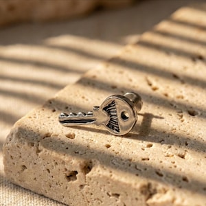 May include: A silver-colored key-shaped earring stud. The earring has a detailed key design with a circular head and a textured surface. The earring is set against a neutral, textured background.
