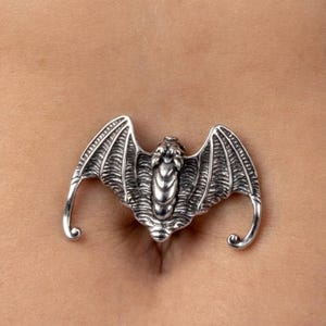 May include: A silver-colored belly button ring in the shape of a bat. The bat has detailed wings and a textured body. The wings curve outwards with a hook at the end of each wing.