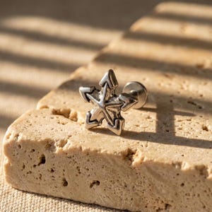 May include: A silver-colored star-shaped earring with arrow-like points. The earring is a stud style, with a flat back and a post for piercing. The star is detailed with lines and curves, and is set against a textured, light brown surface.