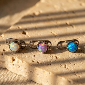 May include: Three dermal anchor jewelry pieces with iridescent opal-like stones. Each piece features a small, round stone set in a silver-colored metal base. The stones display a range of colors, including white, pink, and blue.
