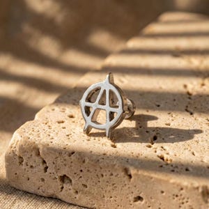 May include: A silver-colored stud earring featuring the anarchy symbol. The earring is a detailed, openwork design with a post backing. The earring is displayed on a textured, light-colored stone surface.
