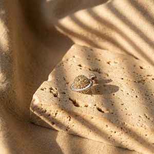 May include: A small, silver-toned stud earring in the shape of a planet. The planet has a ring and is covered in small, yellow-gold colored stones. The earring is resting on a textured, light-colored stone surface.