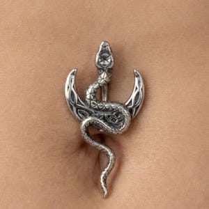 May include: Silver-toned belly button ring featuring a detailed snake coiled around a crescent moon. The snake's scales and the moon's intricate design are visible. The jewelry is designed to be worn as body jewelry.