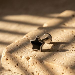 May include: A silver-colored stud earring with a black star-shaped design. The earring is set against a textured, light-colored surface, with shadows adding depth. The star has a defined outline.