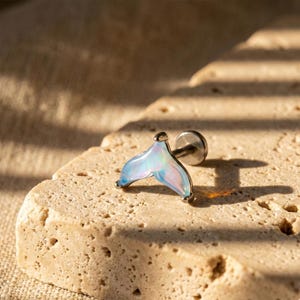 May include: A silver-colored stud earring featuring a mermaid tail design. The tail is iridescent with shades of blue, pink, and white. The earring is set against a textured, light brown surface.