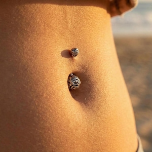 May include: Close-up of a belly button with two silver belly button rings. The top ring is a small ball, and the bottom ring is a larger, ornate design. The skin is tanned and smooth, with a blurred background.