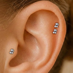 May include: Close-up of an ear with three small, silver cherry stud earrings. Each earring has two round, white pearls and a small, leaf-shaped design with clear crystals. The earrings are placed on the ear's cartilage.