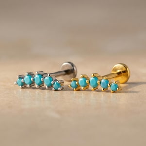 May include: Two ear piercing studs. One is silver-colored, the other is gold-colored. Each stud features a row of five turquoise-colored gemstones. The studs are displayed on a light brown surface.