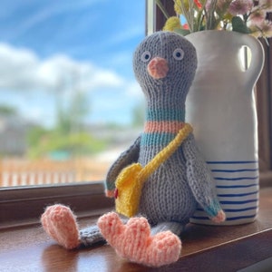 May include: A grey knitted bird toy with a yellow satchel and pink feet. The bird has large white eyes and a pink beak. The bird is sitting on a wooden surface next to a white vase with blue stripes.