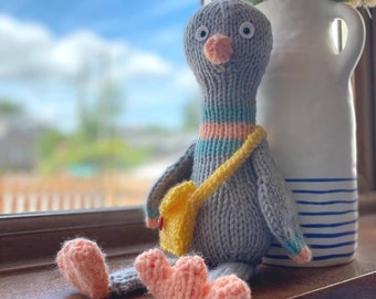 May include: A gray knitted bird toy with a yellow satchel and pink feet. The bird has large white eyes and a pink beak. The bird is sitting on a wooden surface next to a white vase with blue stripes.