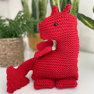 May include: A red knitted dragon toy with black eyes. The dragon is sitting on a white surface.