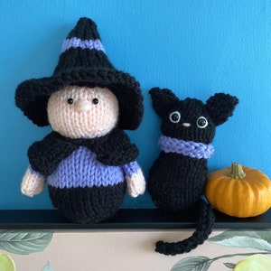 May include: A black knitted witch doll with a purple hat and a black knitted cat doll with a purple scarf. The dolls are sitting on a shelf in front of a blue wall.