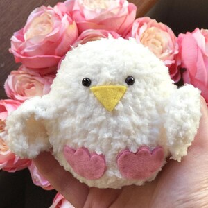 Puede incluir: Un juguete de peluche de pollito blanco y esponjoso con ojos negros, pico amarillo y patas de fieltro rosa. El pollito está sentado en una mano rodeado de rosas rosas.