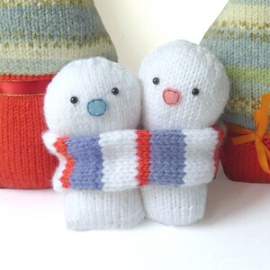 Könnte beinhalten: Zwei weiße gestrickte Schneemann-Ornamente mit rot-blau gestreiften Schals. Der Schneemann links hat eine blaue Knopfnase und schwarze Augen. Der Schneemann rechts hat eine rosa Knopfnase und schwarze Augen.