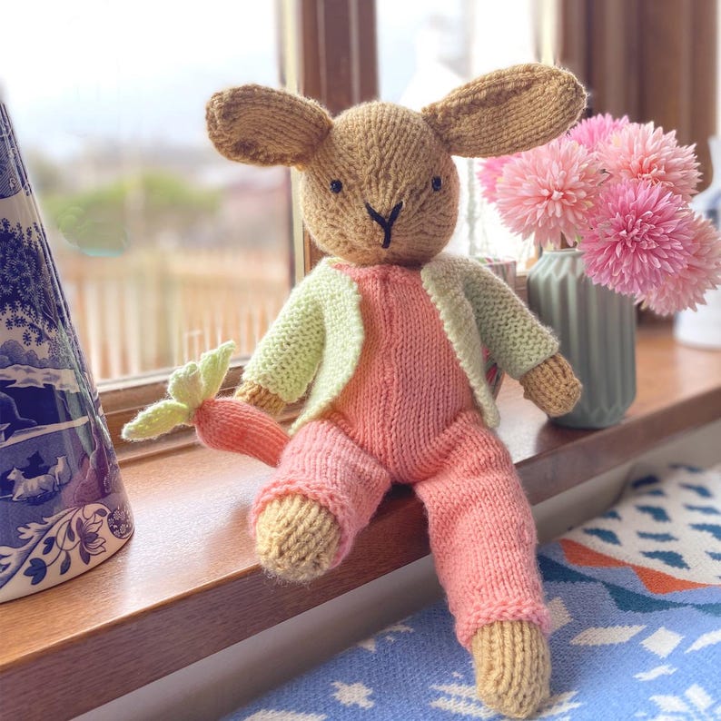 May include: A knitted bunny toy wearing a pink jumpsuit and a light green cardigan. The bunny is sitting on a windowsill with a bouquet of pink flowers in a vase behind it.