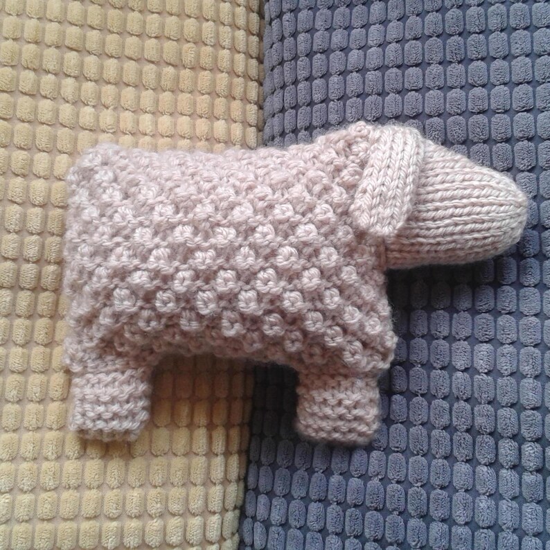May include: A knitted pink sheep toy with a textured surface. The toy is sitting on a yellow and blue patterned fabric.
