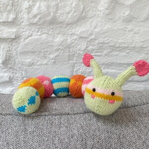 Könnte beinhalten: Ein gestricktes Raupenspielzeug mit einem hellgrünen Kopf und Körpersegmenten in verschiedenen Farben. Der Kopf hat rosa Spitzenantennen und ein gestreiftes Design. Die Körpersegmente sind orange, rosa, blau und gelb mit verschiedenen Streifenmustern.