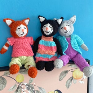 Könnte beinhalten: Drei gestrickte Katzenspielzeuge. Eines ist orange mit rosa und grüner Kleidung, eines ist schwarz mit einem rosa-blau gestreiften Kleid und eines ist grau mit lila Hosen und einer hellblauen Strickjacke.