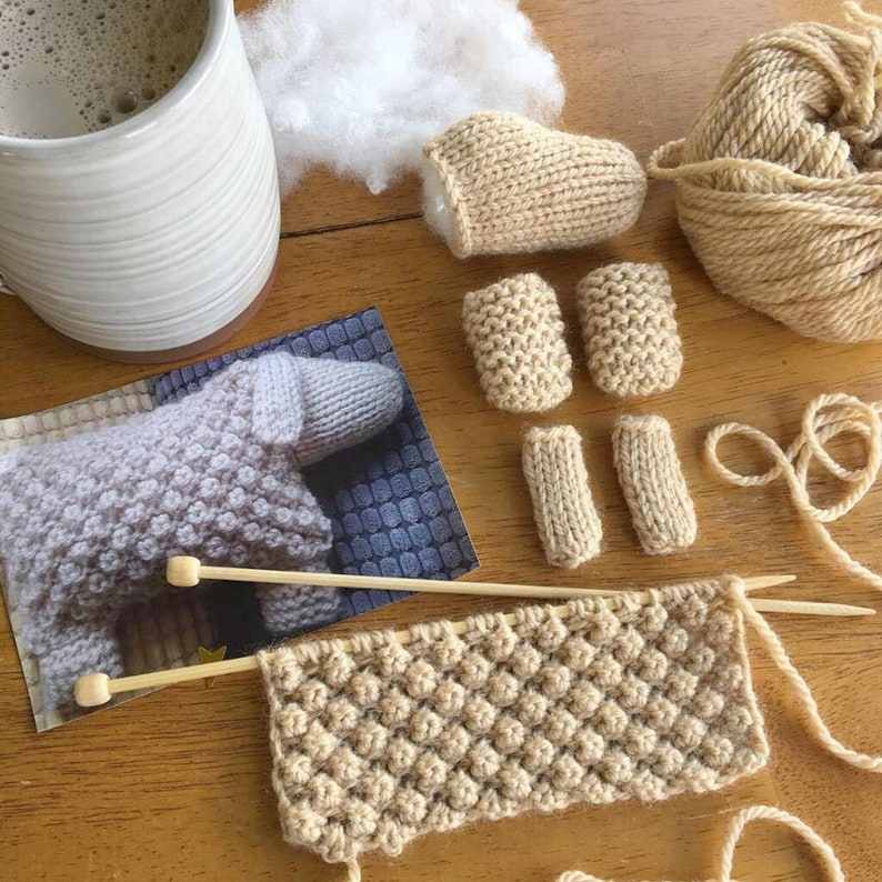 May include: A close-up of a knitting project with beige yarn. The project is a small knitted animal, with pieces for the body, legs, and head. There is also a ball of yarn, knitting needles, and stuffing for the animal.