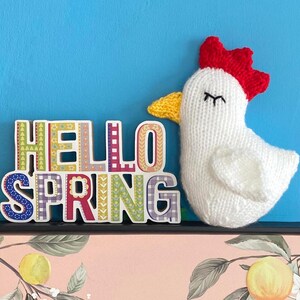 May include: A white knitted chicken with a red comb and yellow beak, next to a colorful wooden sign that reads "Hello Spring".
