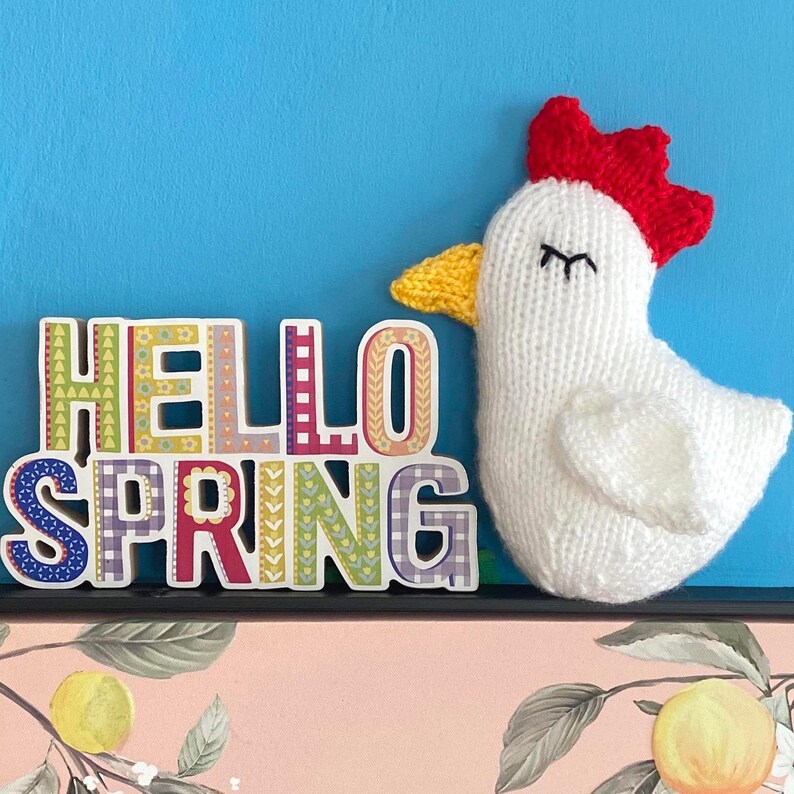 May include: A white knitted chicken with a red comb and yellow beak, next to a colorful wooden sign that reads "Hello Spring".